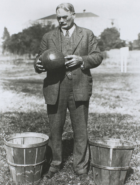 James Naismith kirolaren sortzailea, lehen partida hartan erabilitako futbol baloia eskuetan eta mertxikak biltzeko saskiak albo banatan. (Arg.: Sports Antiques)