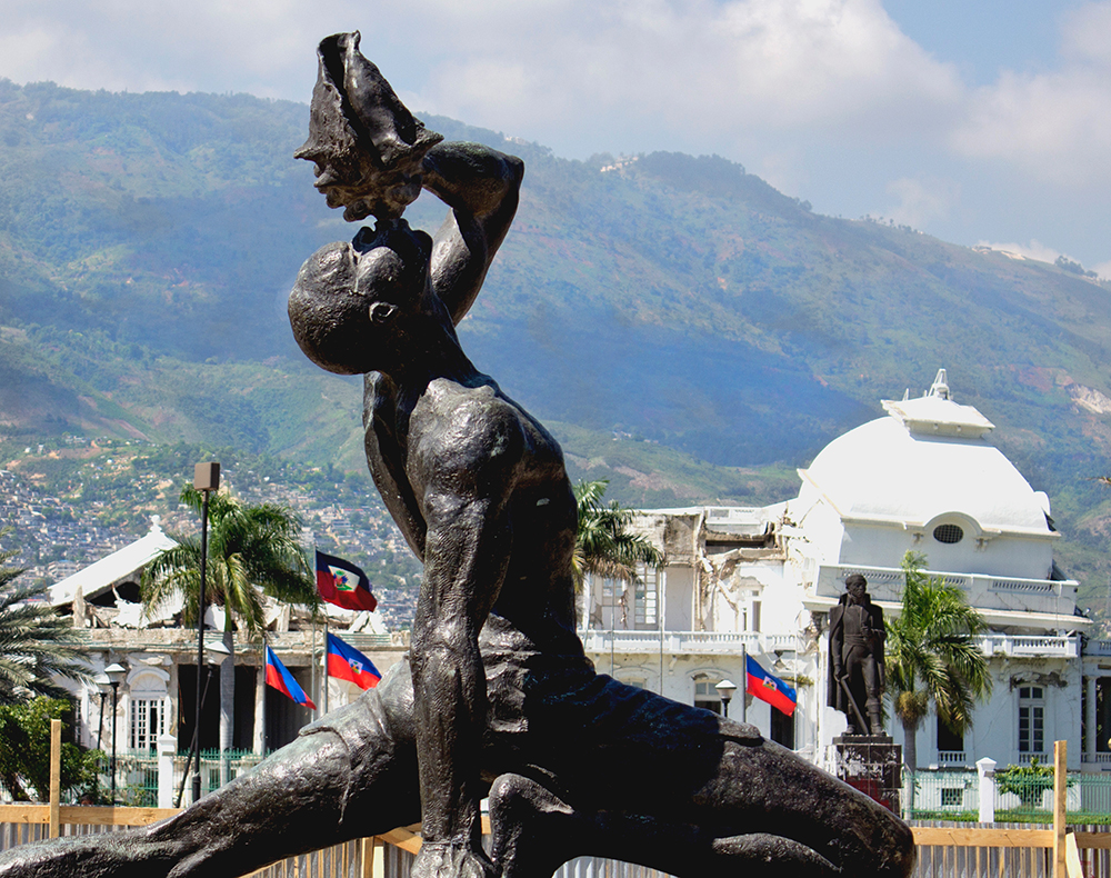 'Le Marron Inconnu' (Esklabo ezezaguna) eskultura Port-au-Princen, esklabotzaren abolizioaren omenez egina. Atzean, eraikin instituzionalak oraindik 2010eko lurrikararen zauriak dituztela. (Argazkia: Wikimedia / Kristina Just)